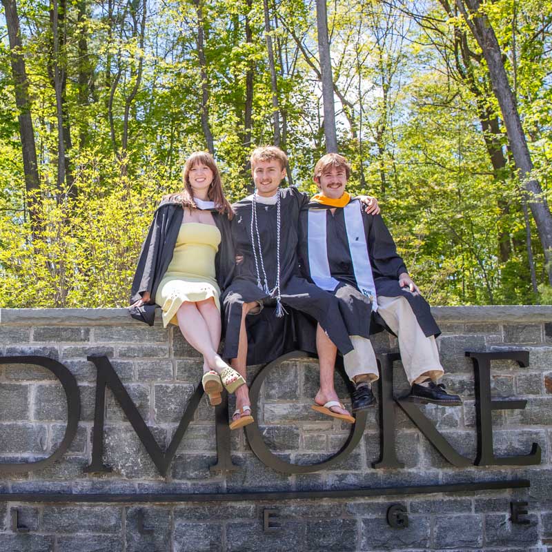 Three graduates in caps and gowns sit together on a stone Skidmore sign in a wooded campus setting.