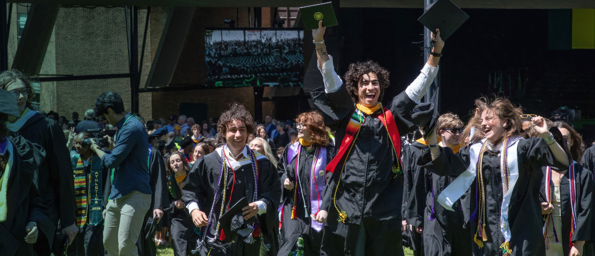 Skidmore Colelge graduates in caps and gowns celebrate outdoors, raising diploma covers as they process from the ceremony.