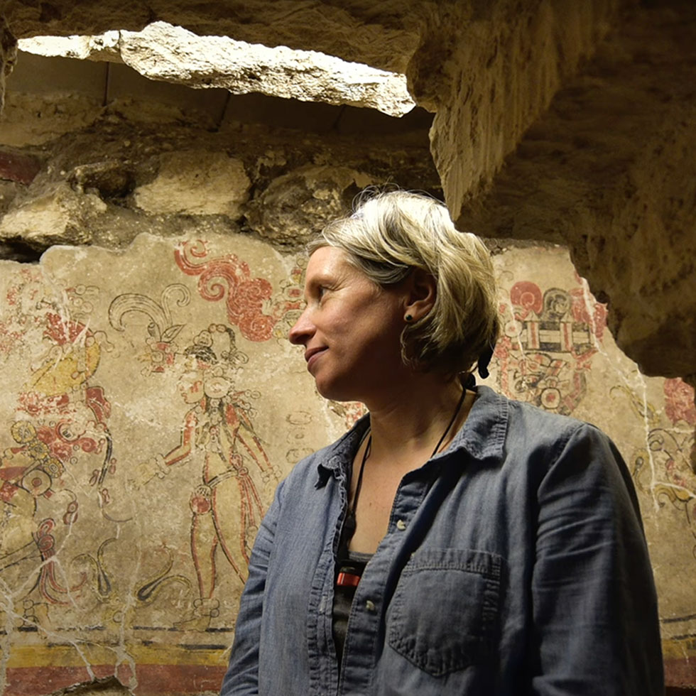 Woman standing in an ancient stone room with partially restored Mayan murals on the wall behind them, illuminated by natural light from an opening above.