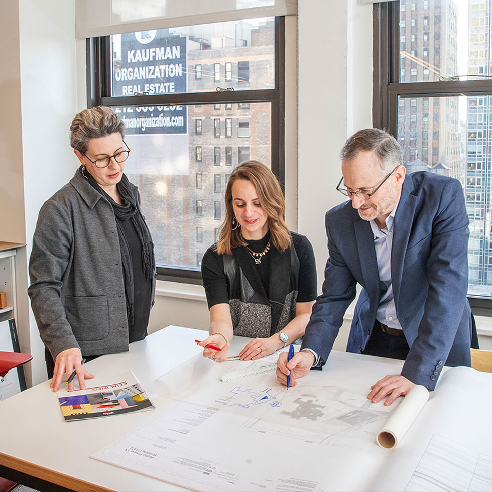 Three professionals collaborating over architectural blueprints in a bright office with large windows overlooking a city skyline.