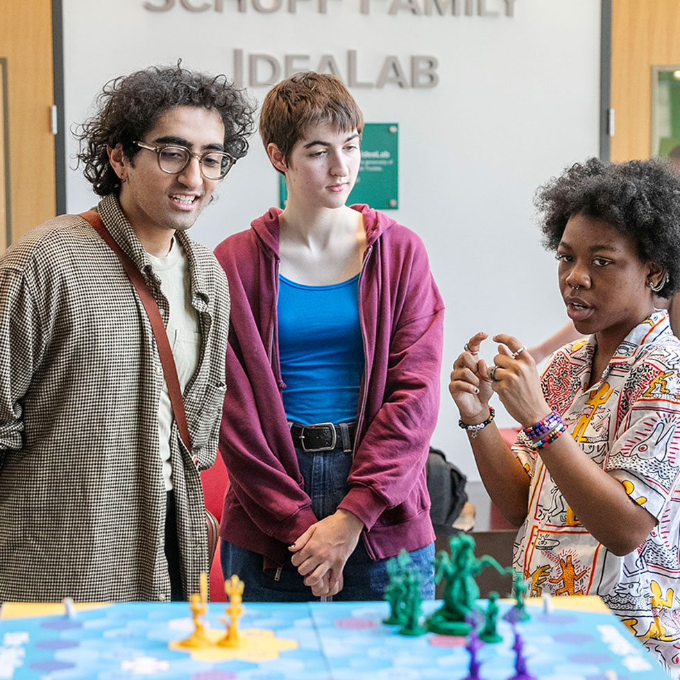 Three students in the Schupf Family IdeaLab engaged in a creative discussion around a colorful tabletop game with custom 3D-printed pieces.