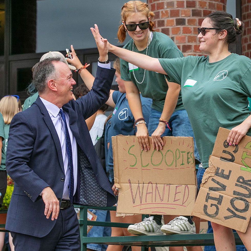 Skidmore College President high-fiving a student during a campus welcome event, surrounded by students holding handmade cardboard signs including one that reads “Scoopies Wanted.”