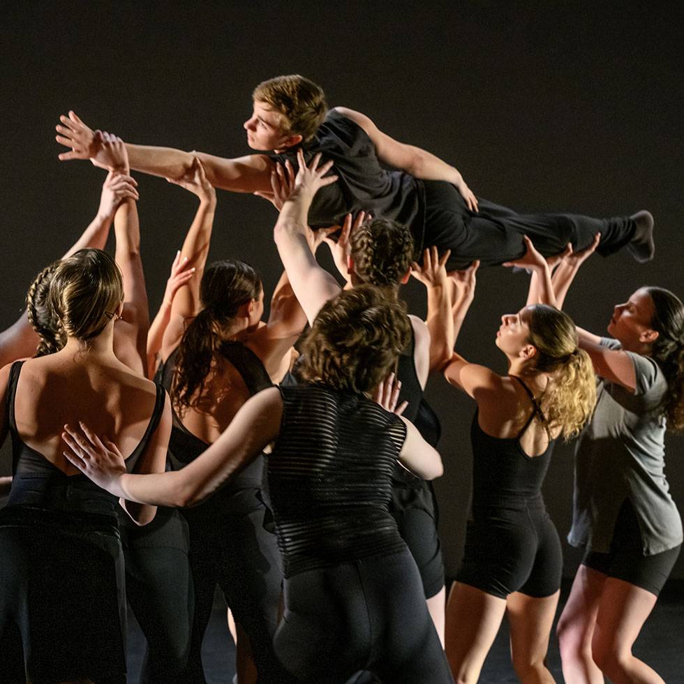 A dark stage is filled with students performing, including a group holding up one dancer laying horizontal with one hand extended over head. All are wearing black.