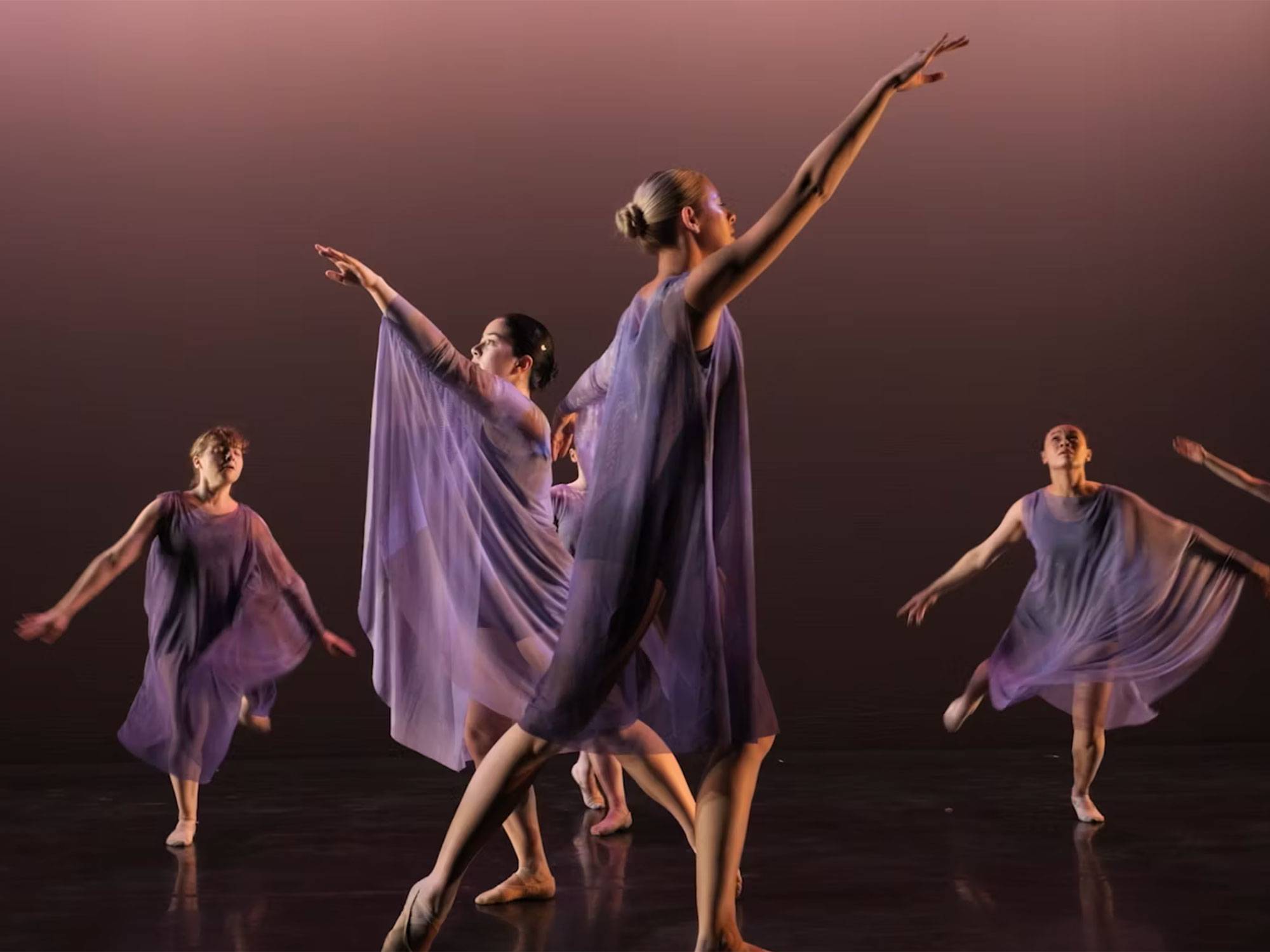 Multiple dancers perform on stage wearing flowing purple costumes