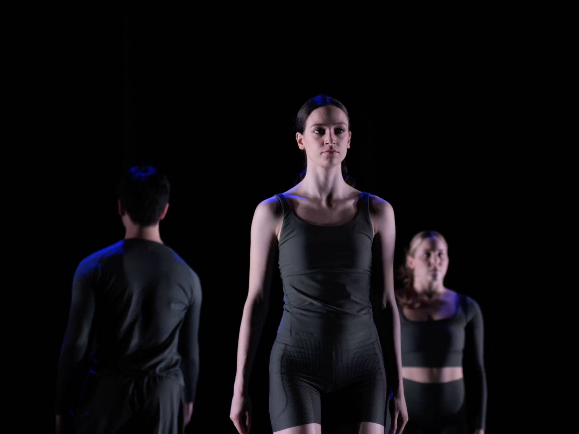 Dancer stands on stage waiting to begin routine, with an all black background and and other dances in black costumes.