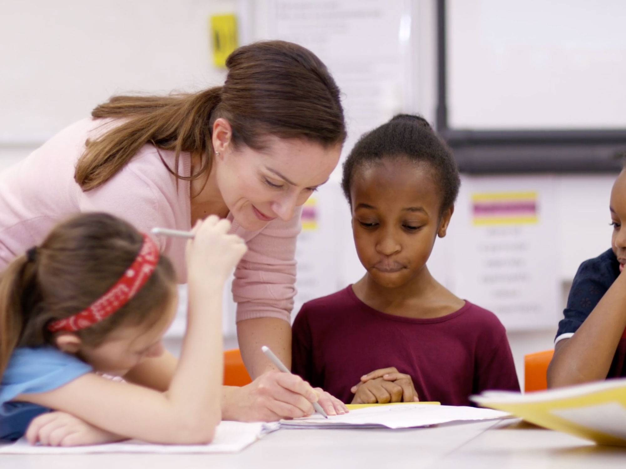 A teacher leans over a table helping students. She holds a pen and is writing on a piece of paper.