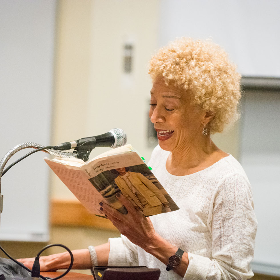 An author stands at a podium, speaking into a microphone with her book in hand reading and smiling