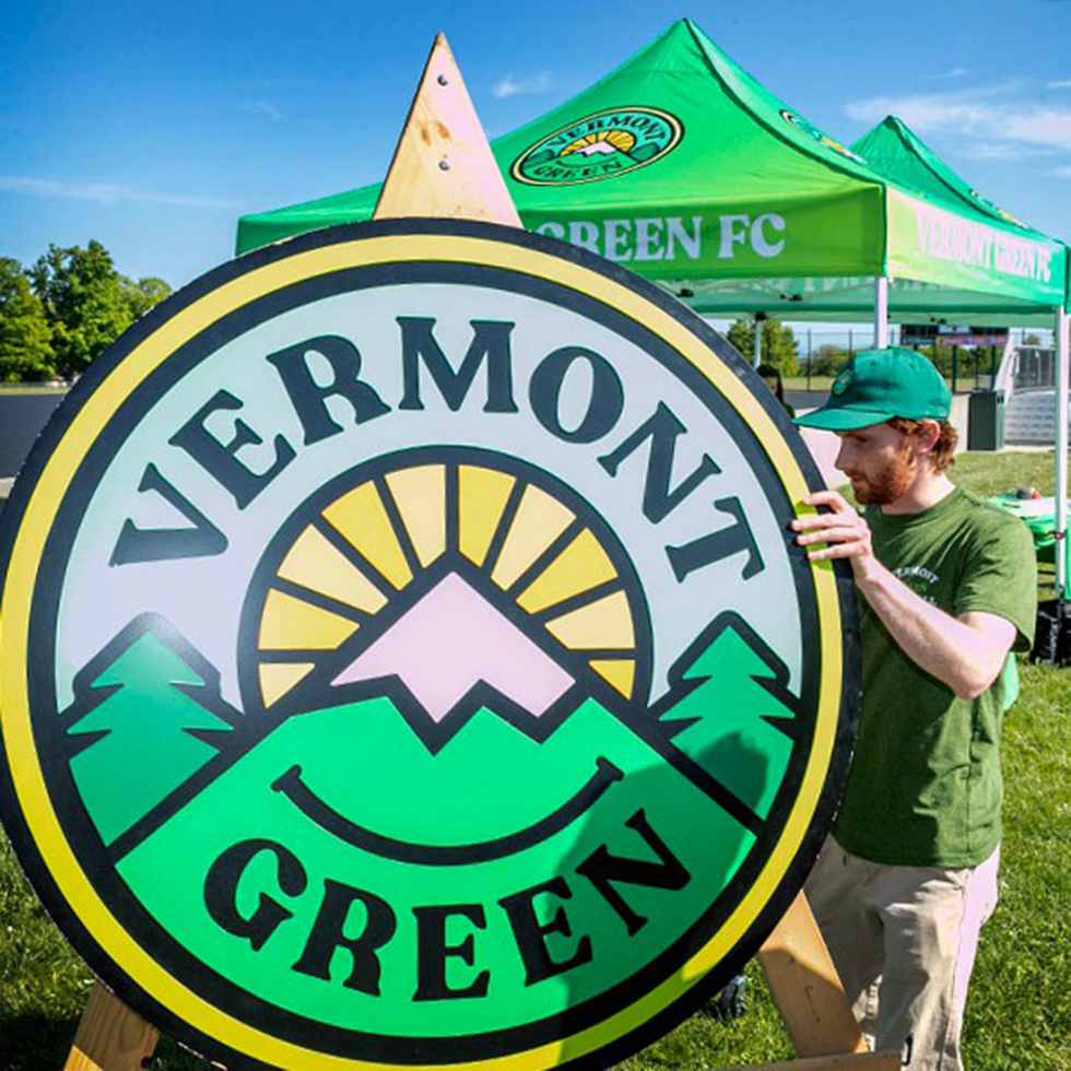 Man in green shirt and baseball cap stands beside a sign with the Vermont Green logo on a sunny day.