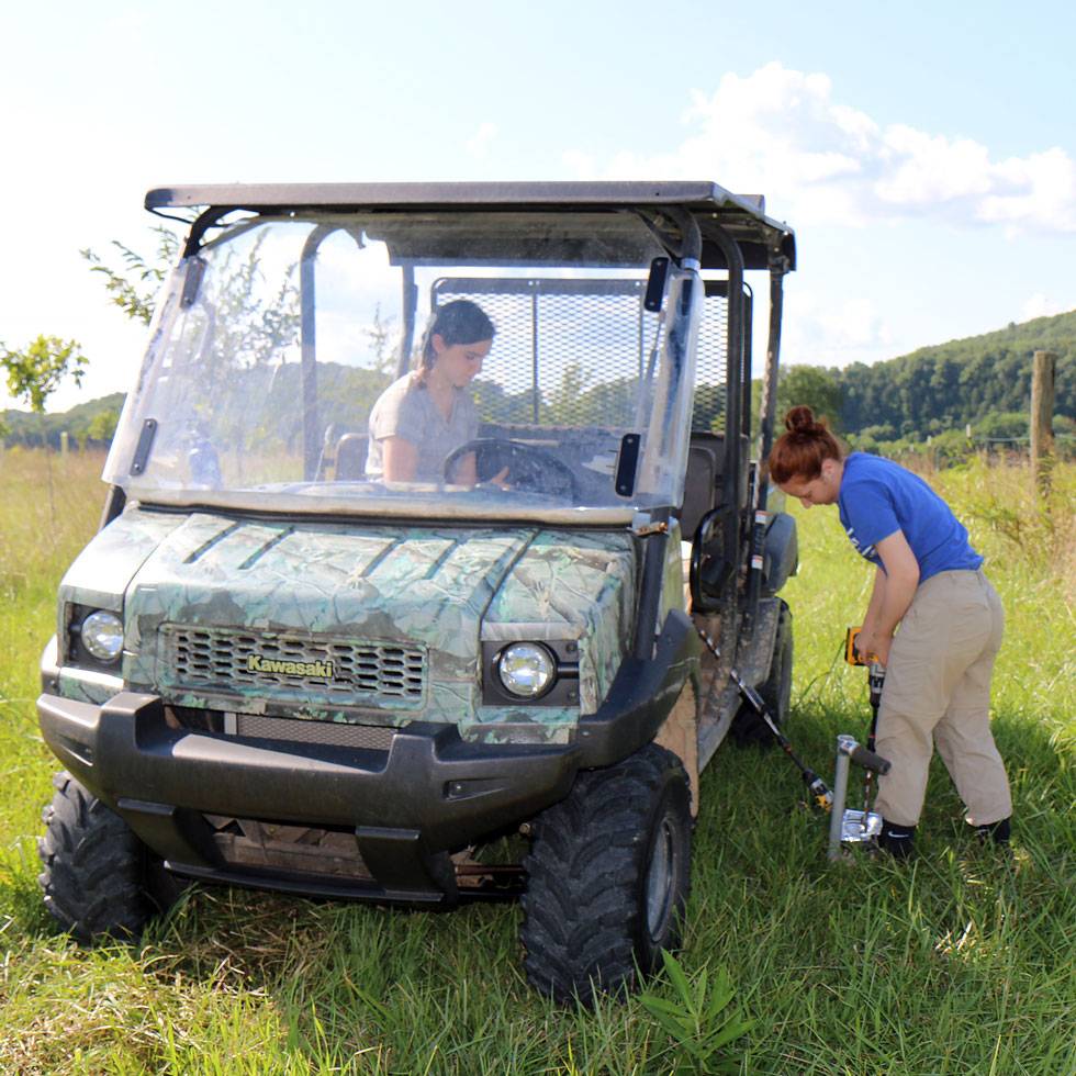 Two students work in a green field with various tools to research soil, including a 4x4 vehicle