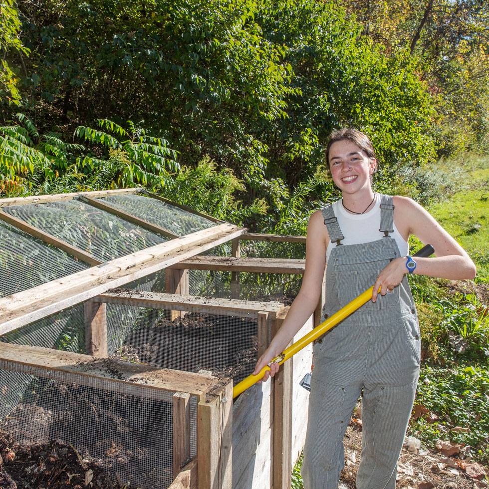 A student smiles in overalls while holding a gardening tool next to a large compost bin made out of mesh wire and a wooden frame.
