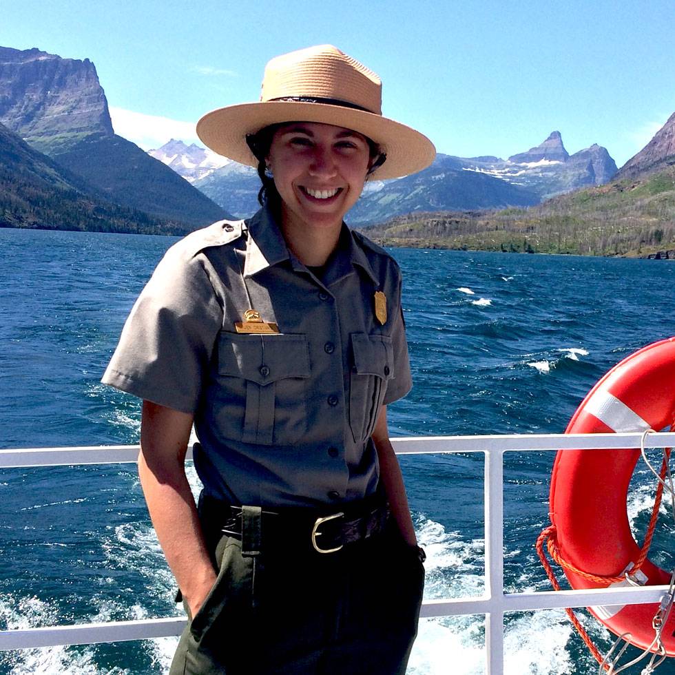 Jennifer Cristiano '18, a park ranger, stands on a boat with a lake and mountain peaks in the background, smiling during a field experience in a national park setting.