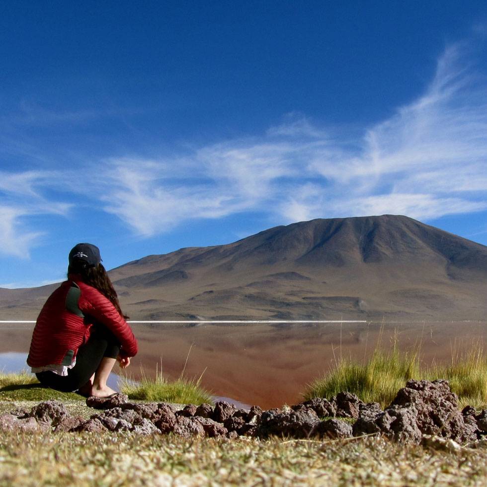 A student crouches down in a red jacket and black hat looking out over a lake and mountain with a blue sky in the background in Bolivia.