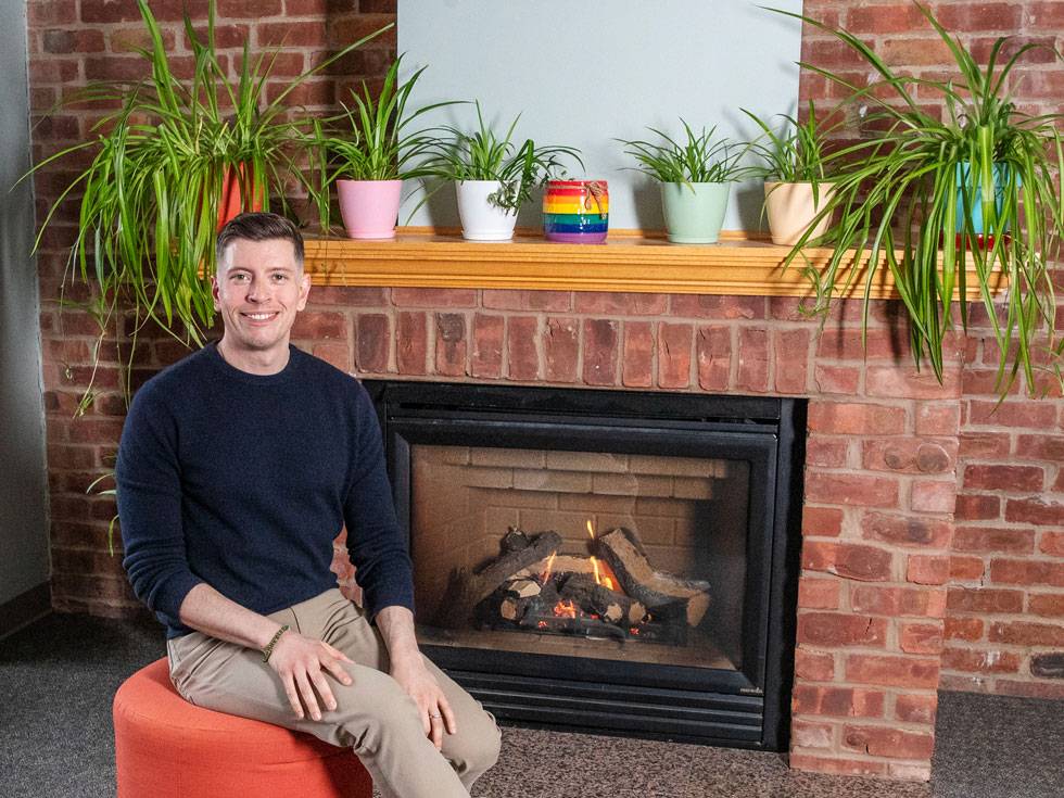A person sits smiling on a bright orange pouf with a brick fireplace in the background and green plants lining the mantel. 