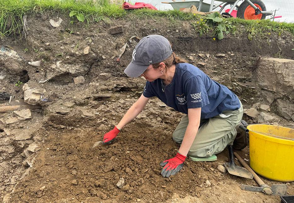 A student is on hand's and knees in a dig site, surrounded by stone walls, tools, and partially uncovered earth.