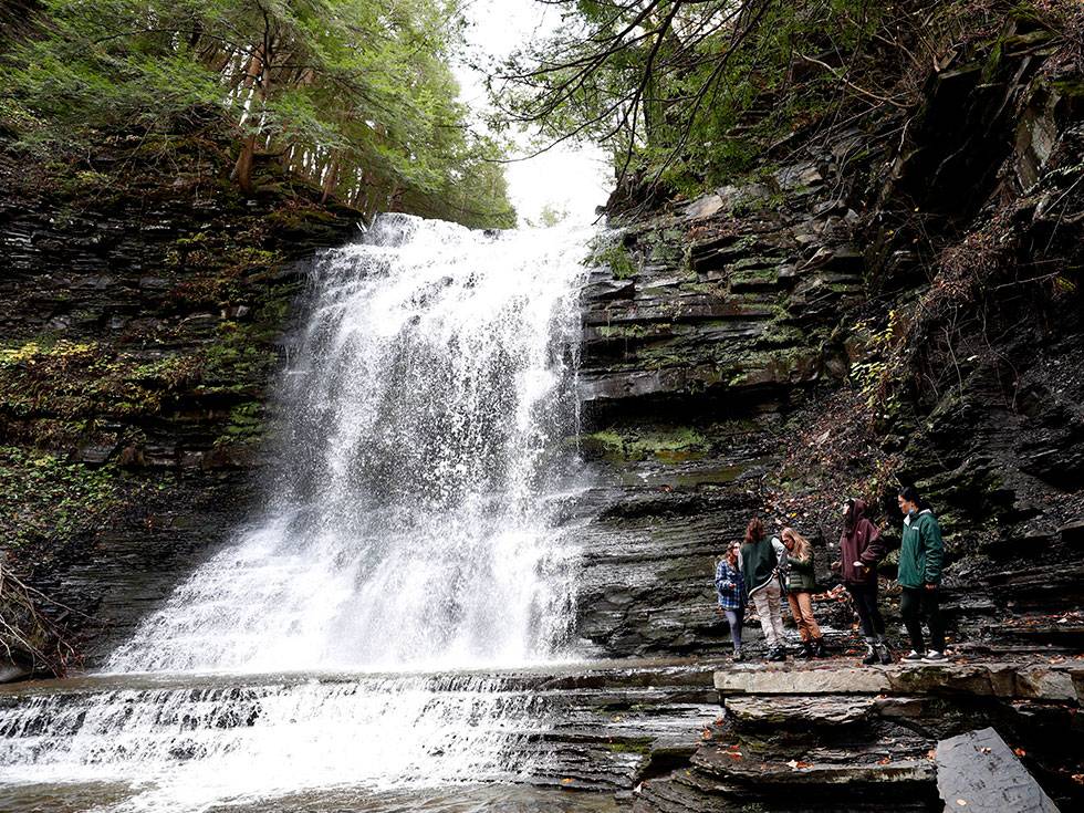 A group of students stand to the right of a waterfall with layered rocks and green moss surrounding