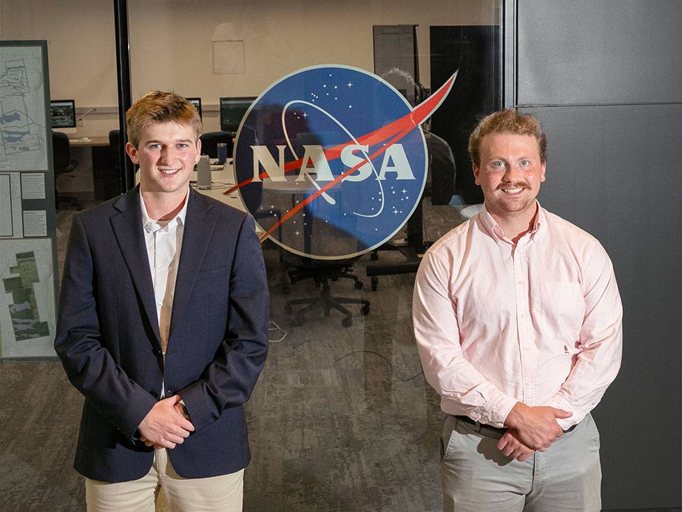 Two individuals pose in front of a large NASA logo inside an office setting. One is wearing a dark suit and white shirt, and the other is wearing a light pink button-down shirt. Computer workstations are visible in the background.