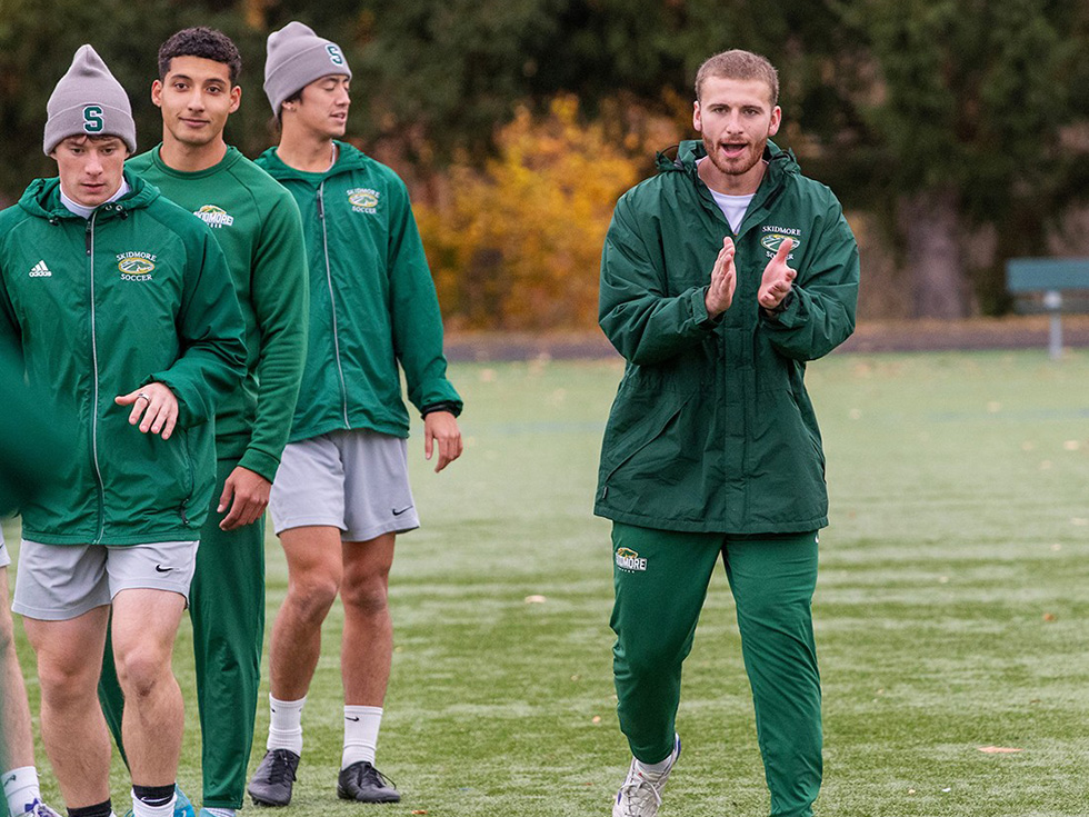 Four members of the Skidmore men’s soccer team walking on a field, with Owen Brady clapping encouragement during practice.
