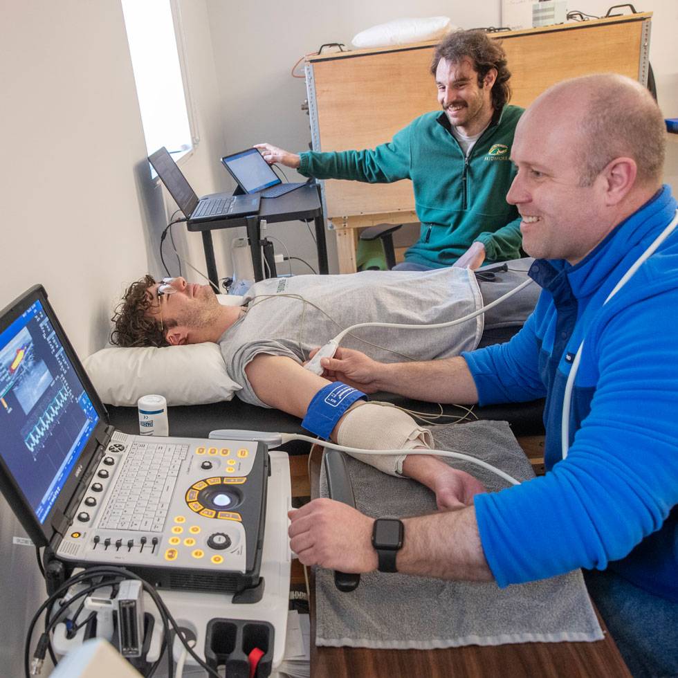 Three individuals work in a lab, gathering health data on the one laying down on a medical bed. 