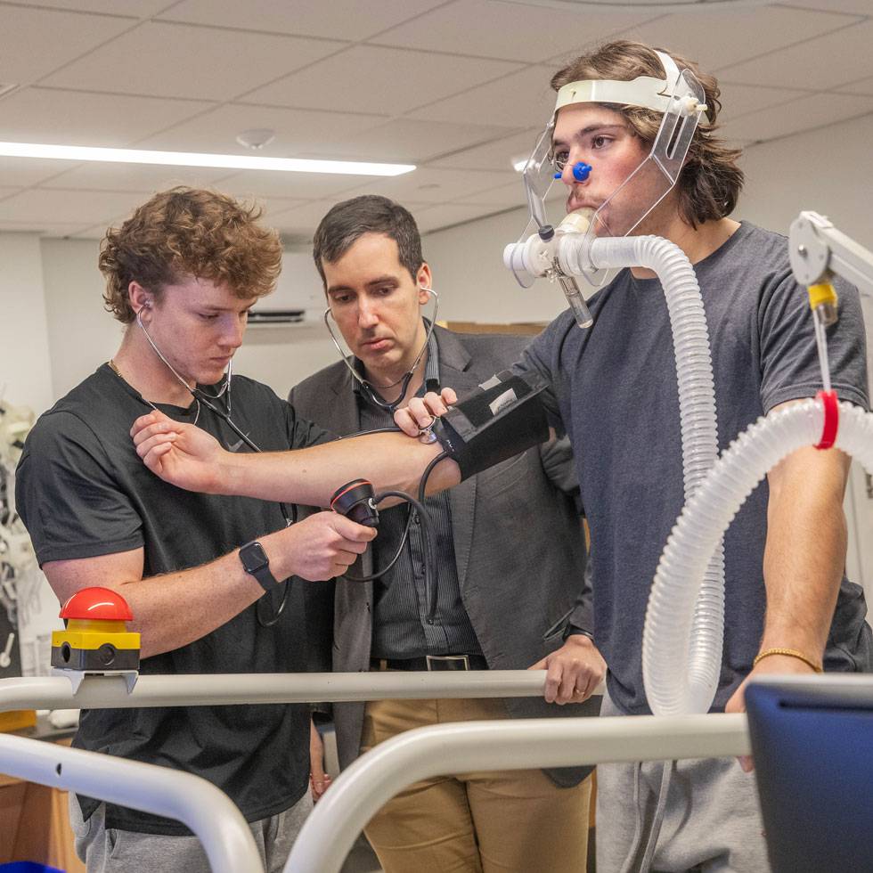 Two students and a professor work in a human performance lab. One is on a treadmill, wearing a mask, with their arm stretched out, while the other measure vitals as a professor looks on.