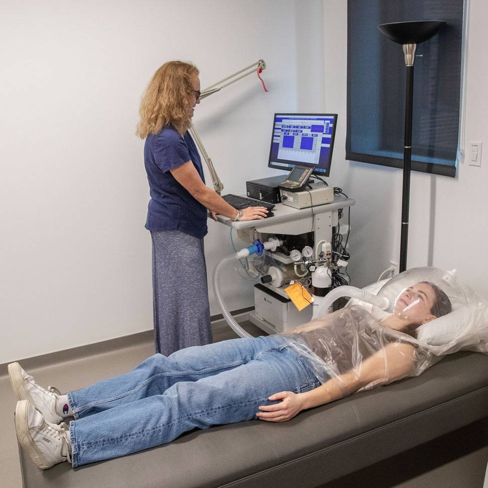 A student lays on a lab bed wearing a clear dome helmet, while a faculty member stands next to them looking at a standing computer screen.