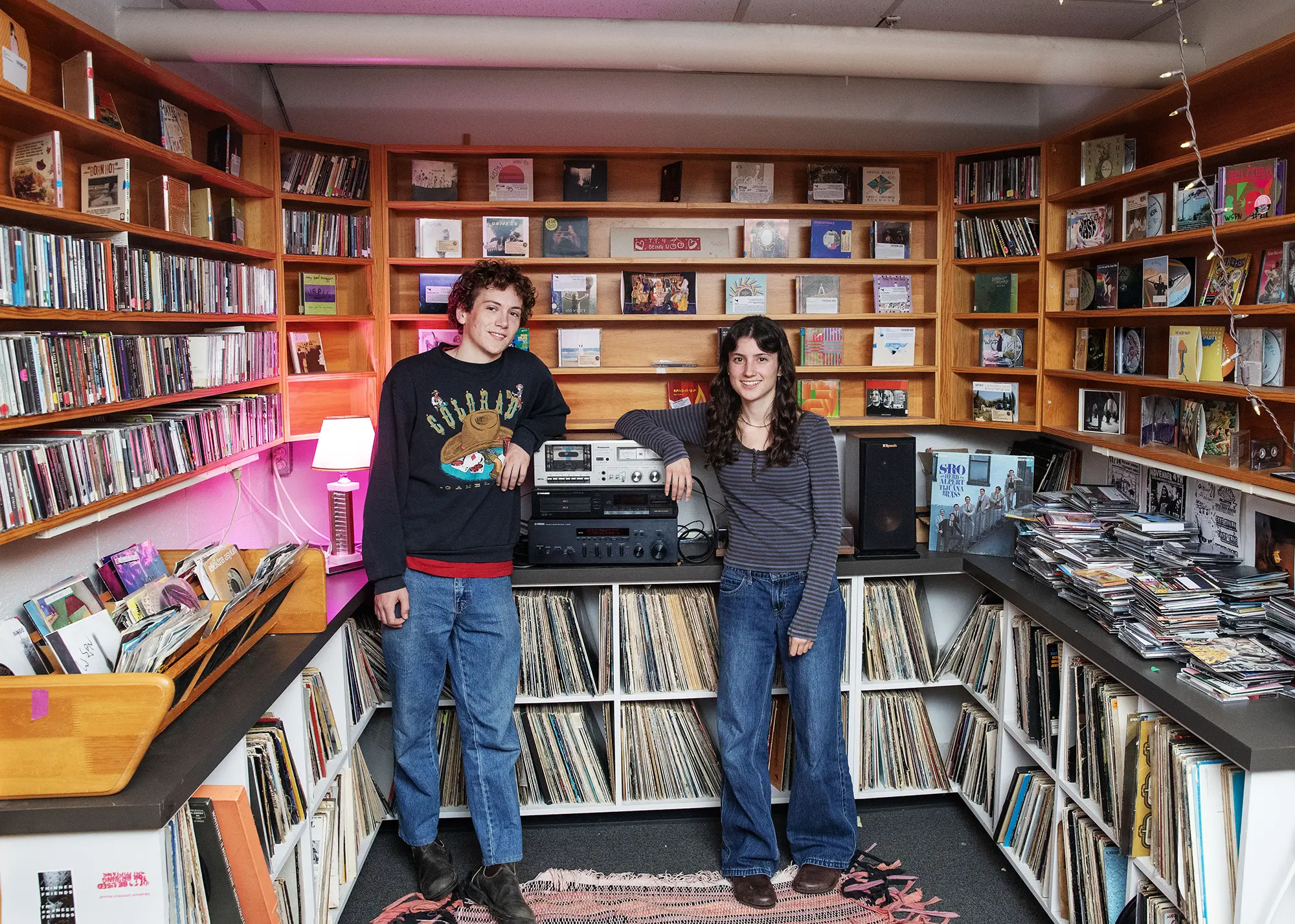 Two students pose in Skidmore College’s WSPN campus radio station, surrounded by shelves filled with vinyl records, CDs, and sound equipment.