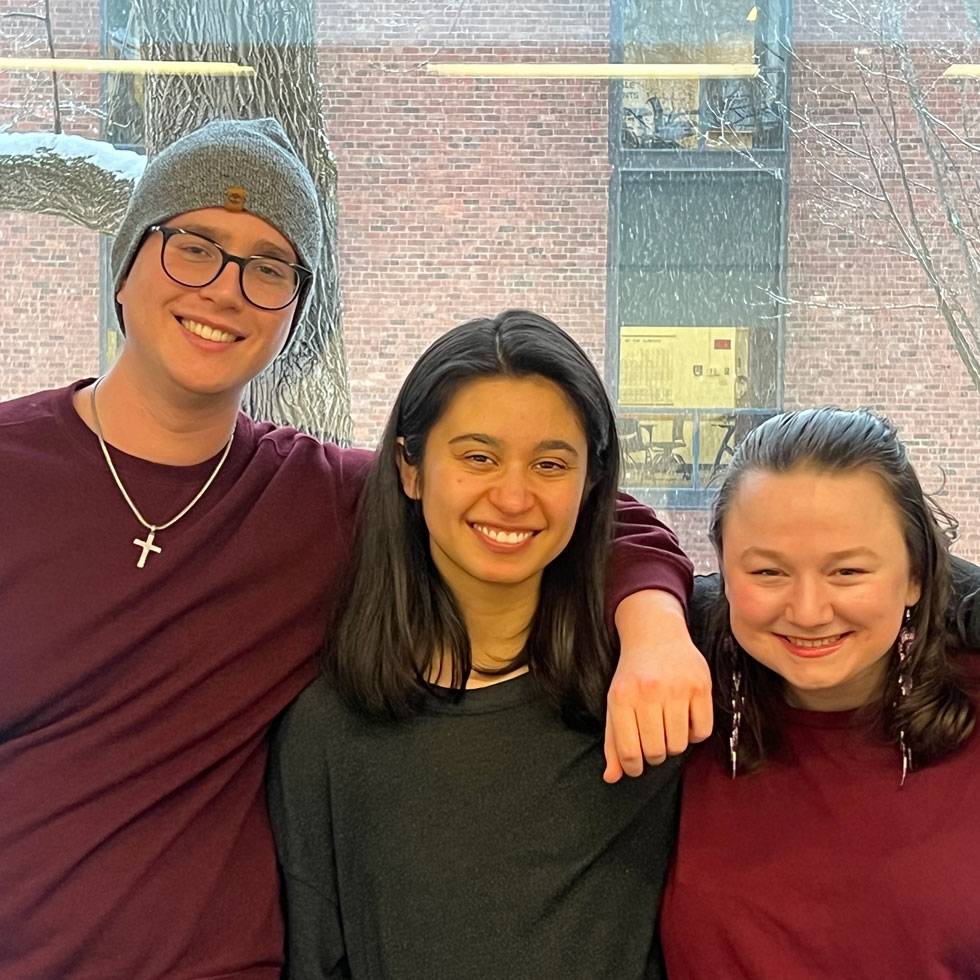 Three students pose in front of a window with a brick building in the background
