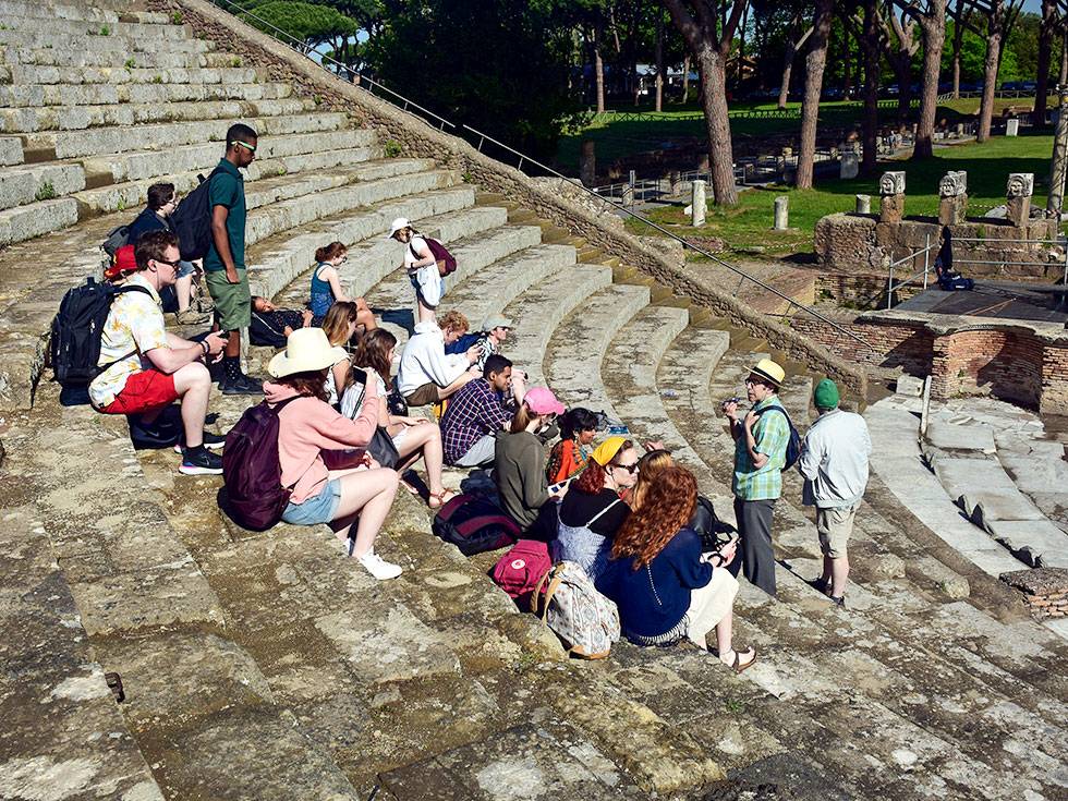 A large group of students sit on classical style steps while a a faculty member speaks to them