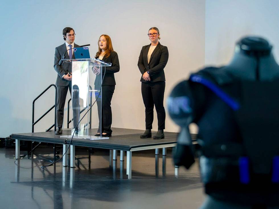Three students stand on stage at podium making a business pitch to an audience. In the right corner foreground, you can see the edge of their product prototype, which is athletic padded equipment in black and purple.