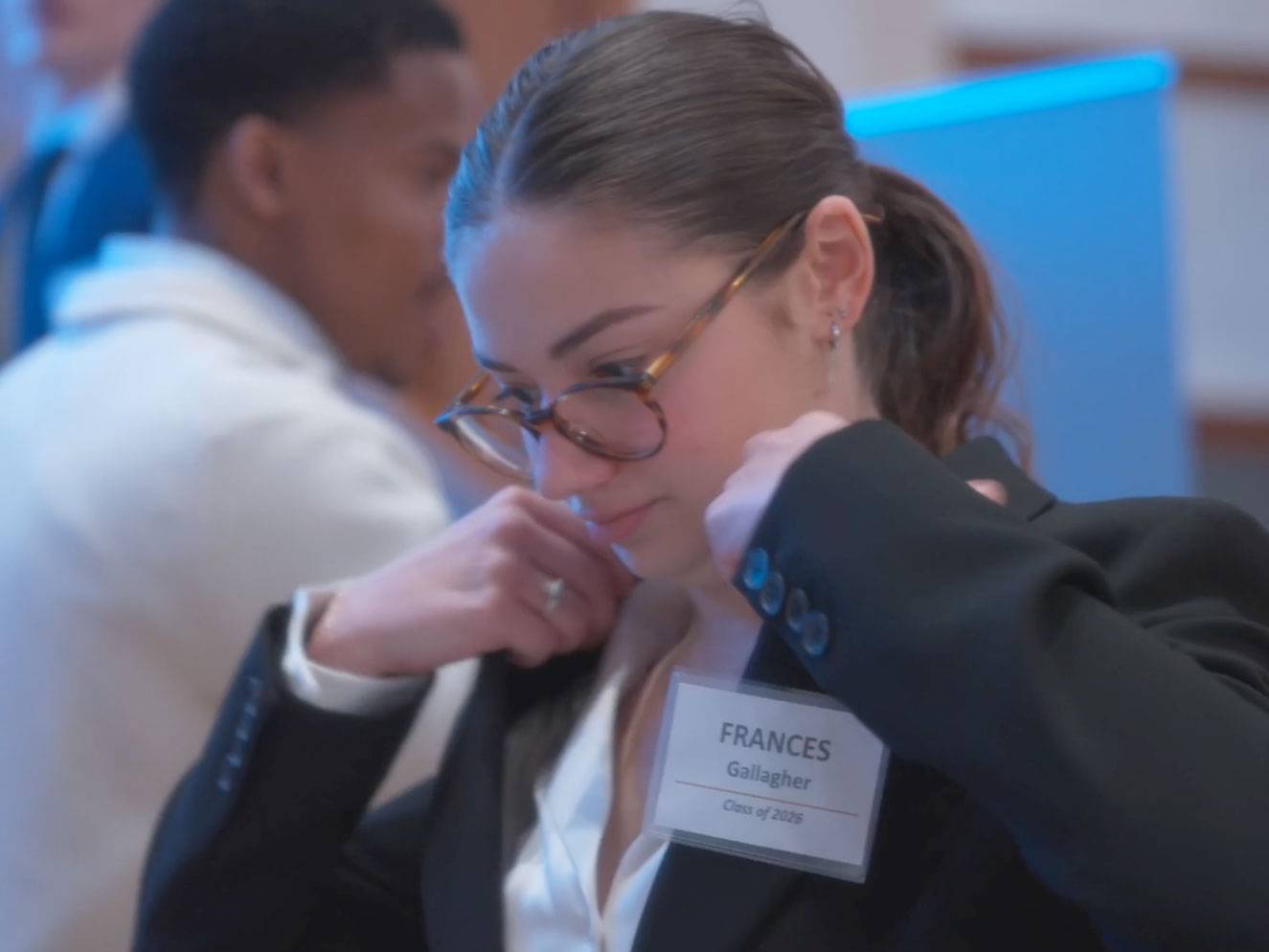 A student fixes the collar of their suit, while wearing a name tag and glasses to prepare for a presentation