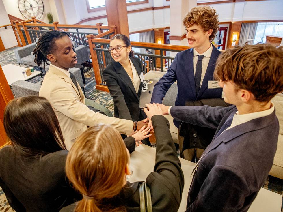 A circle of students dressed in professional suits, all holding their hands in the center of the circle.