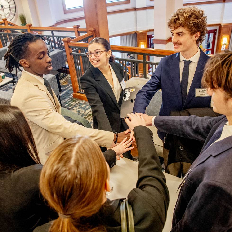 A group of students in business attired stand in a circle with their hands together in the middle smiling as they prepare to present at MB107