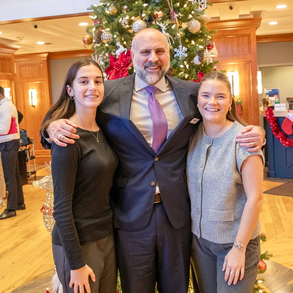 A faculty member stands with his arms around two students on either side. All are wearing business attire, and the background is a hotel lobby.