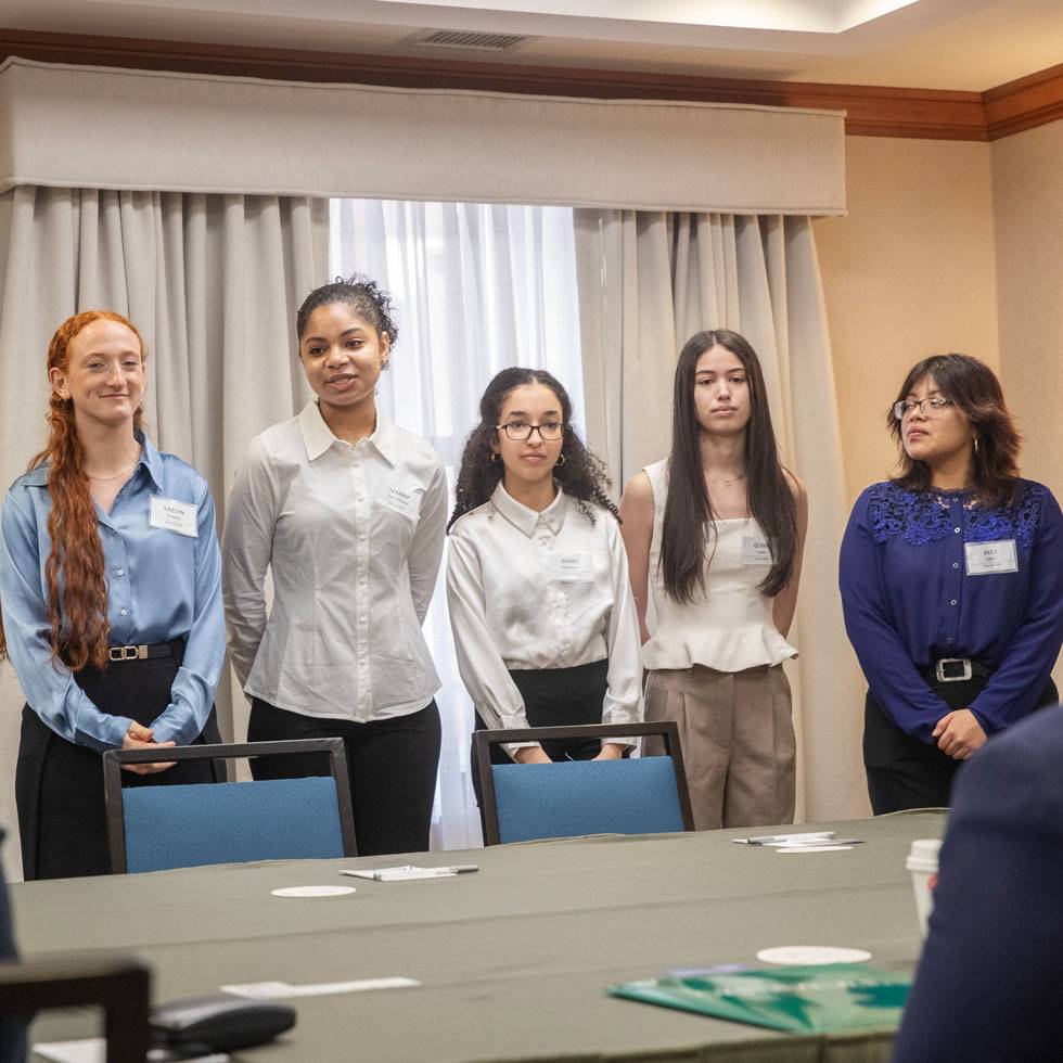 Five students in business attired stand behind a long conference room table preparing to present at MB107