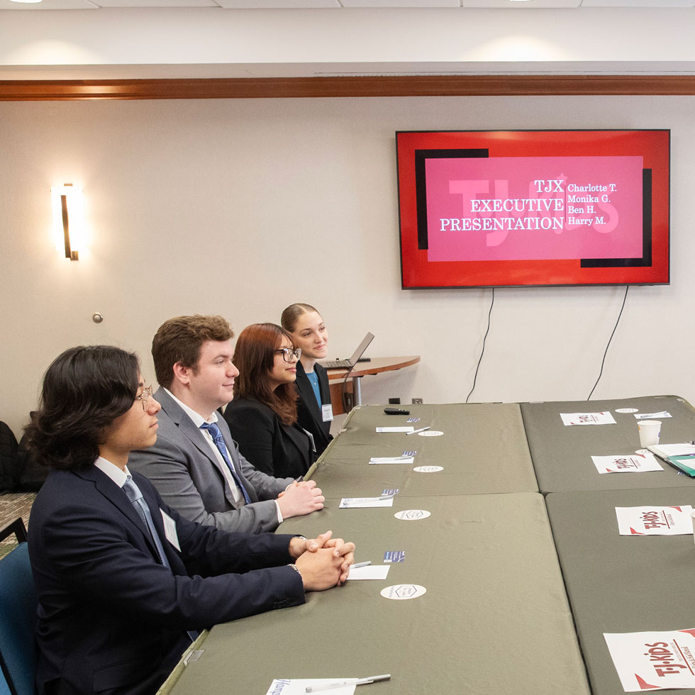Four students sit in business attire at a table, ready to present to executives on the either side of the table (not pictured)