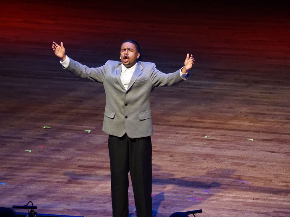 Student on stage singing with raised hands.