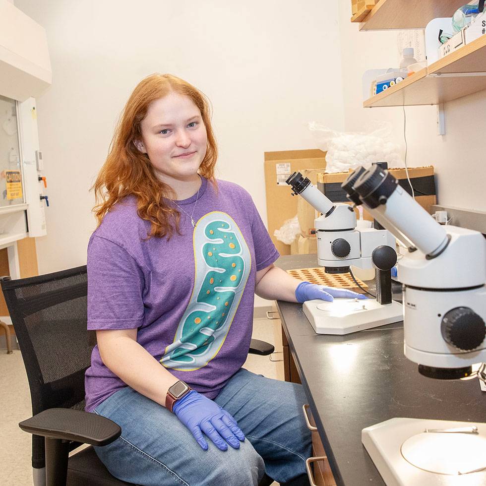 Jordan Diamond '27 sits in a lab, smiling, with a microscope in front of her and wearing a purple shirt and purple lab gloves.
