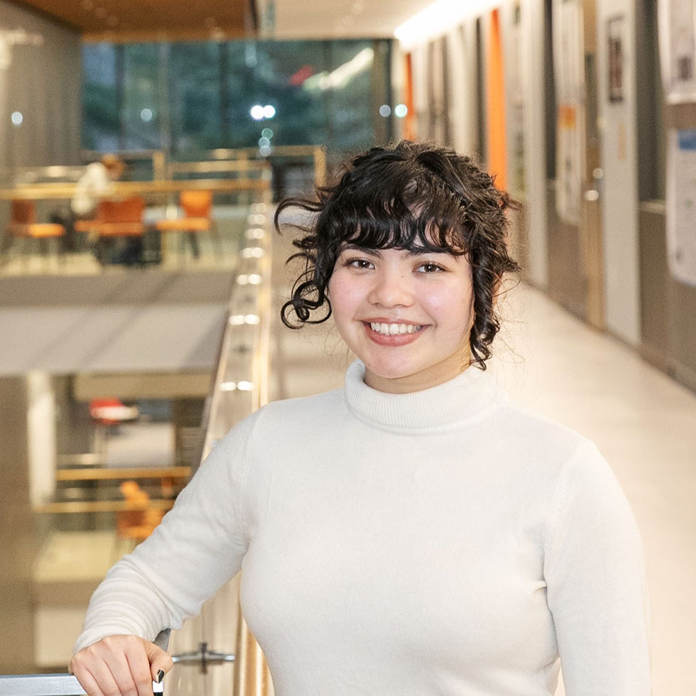 A student in a white turtleneck smiles while standing in a brightly lit academic building hallway.