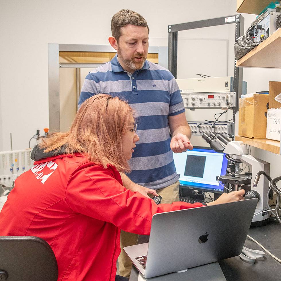 A student sits at a computer with scientific research equipment in the background, along with a professor standing over providing direction..