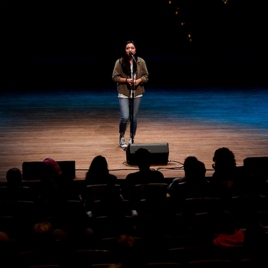 Female+student+on+stage+under+a+spotlight+speaks+into+a+microphone