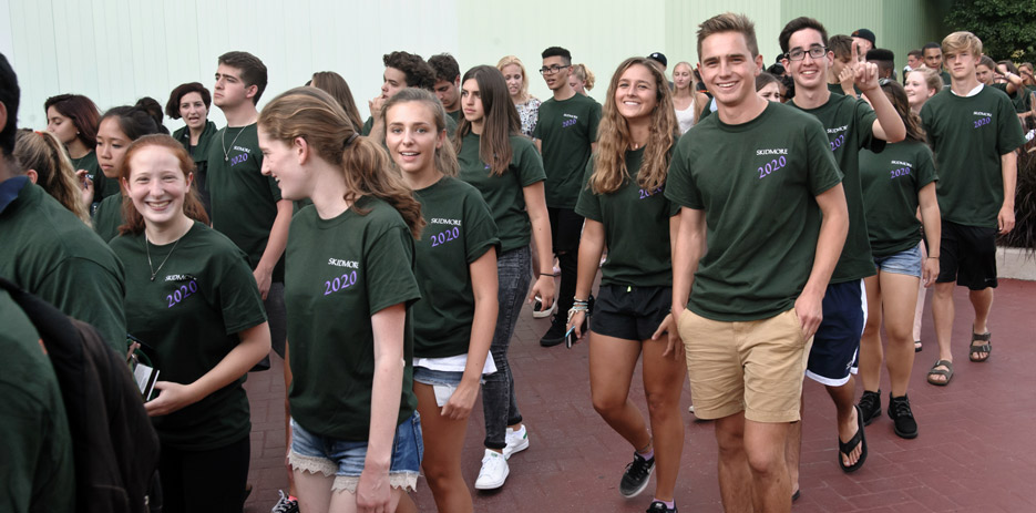 Three college students in matching T-Shirts that say "Skidmore 2020" parade into a ceremony 