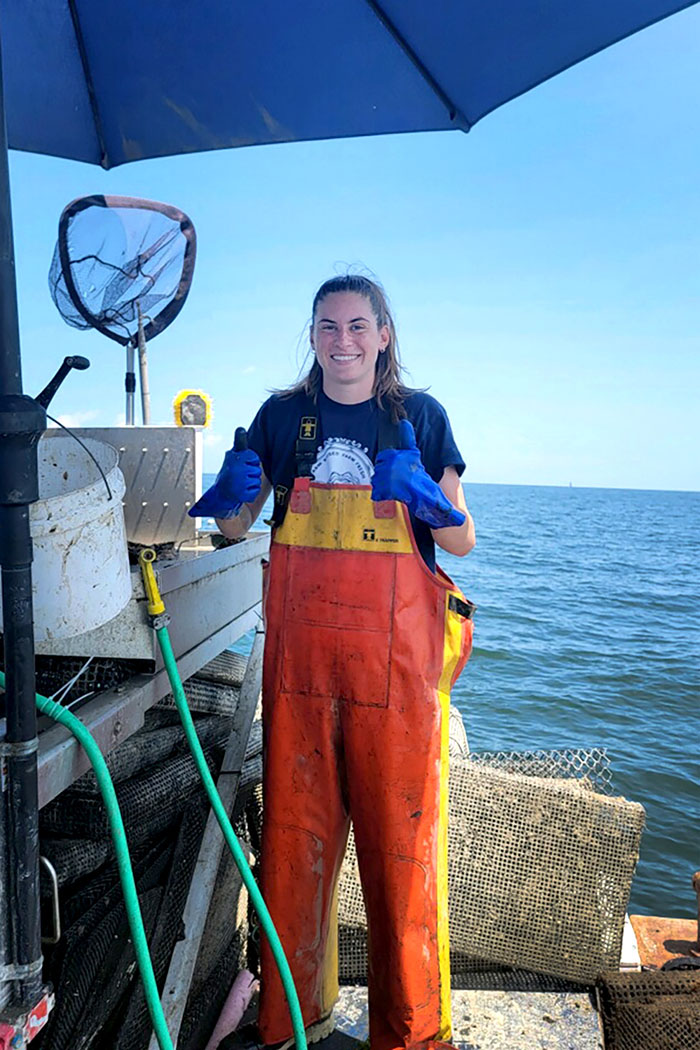 Julia stands on an oyster boat in overalls and gloves, giving two thumbs up and smiling.