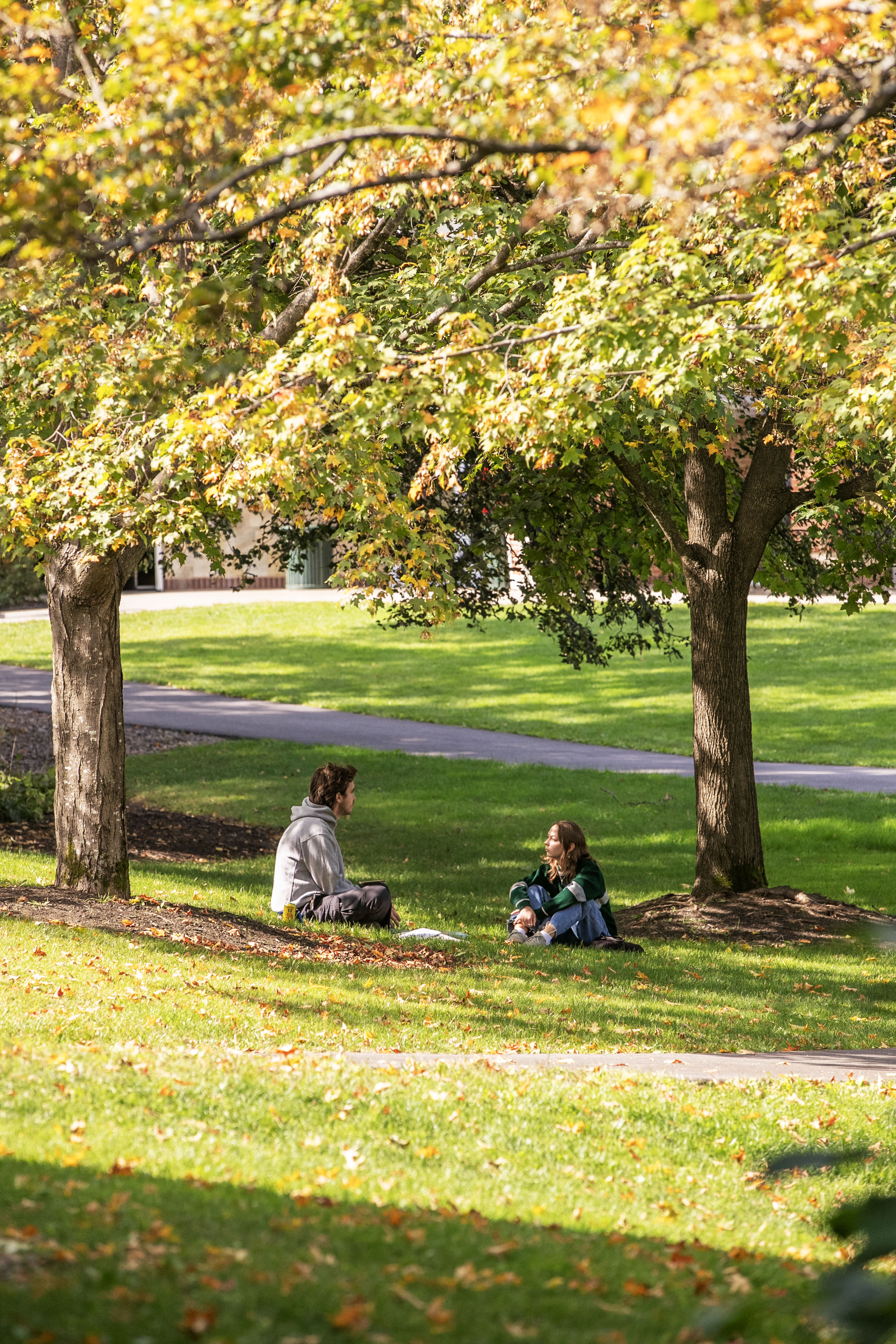 Two students sit on the Skidmore green under fall foliage.