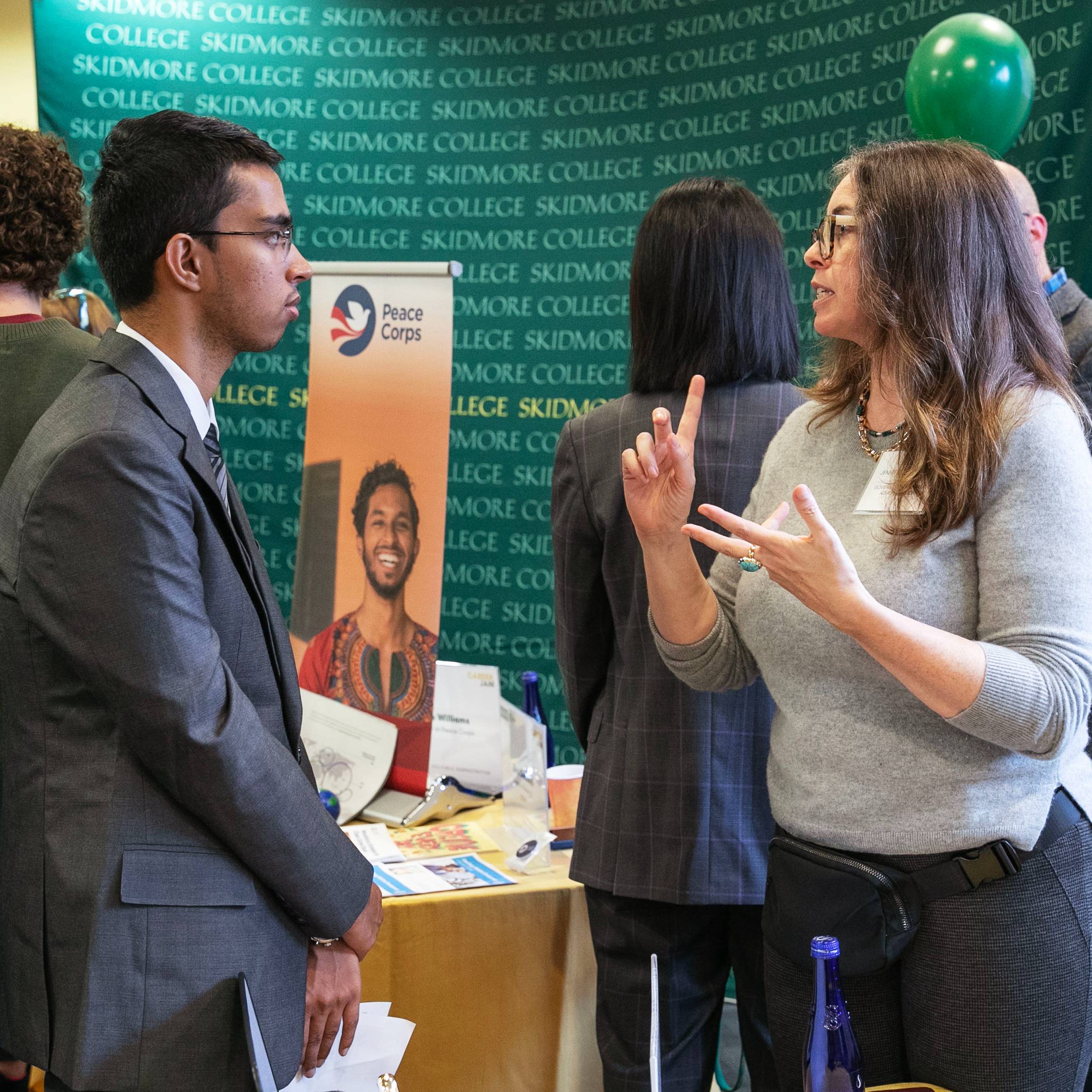 A+Skidmore+student+wearing+a+suit+speaks+to+a+volunteer+mentor.+You+can+see+a+Peace+Corps+sign+and+a+screen+printed+with+the+Skidmore+College+logo+in+the+background.