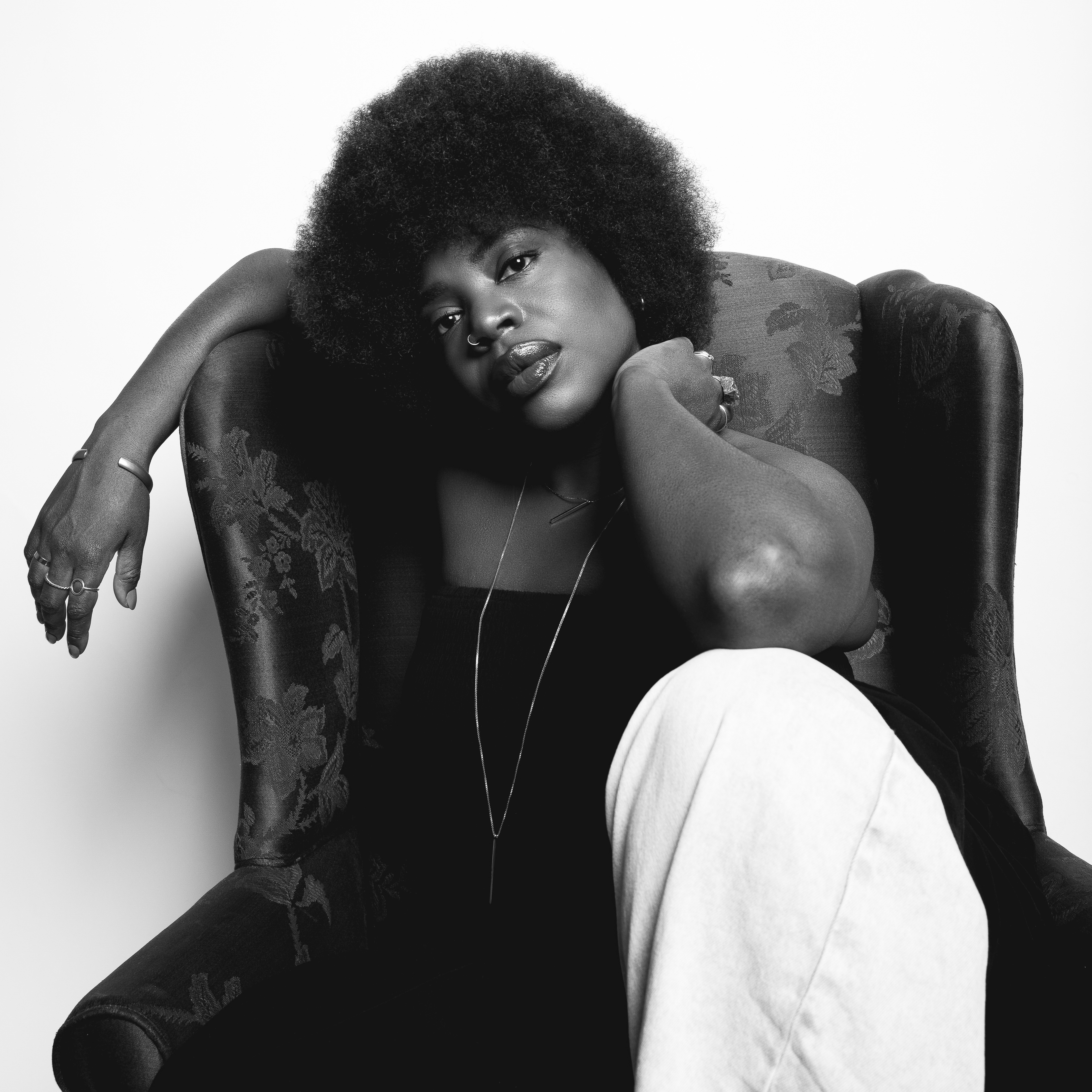 A+black+and+white+photo+of+Melanie+Charles+sitting+in+a+chair.+She+looks+at+the+camera+with+one+knee+propped+up+and+an+arm+resting+off+the+side.