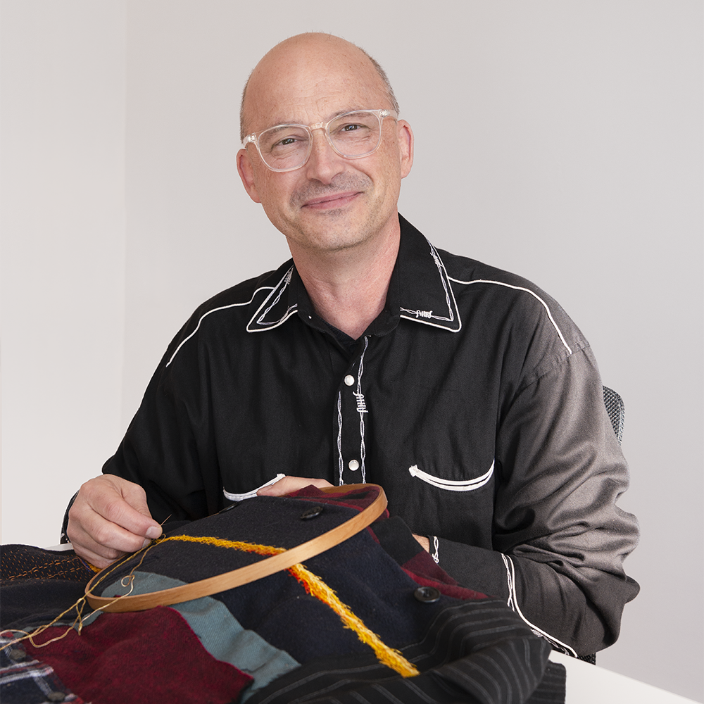 Mark+Newport+looks+up+from+the+project+he+is+embroidering+and+smiles+