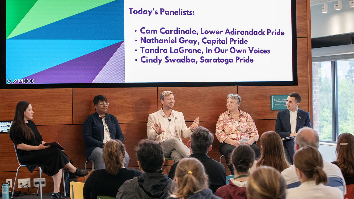 Four panelists and Mariel Martin sit on the Wyckoff Center stage, a bit of the crowd and a large screen introducing each panelist can be seen.