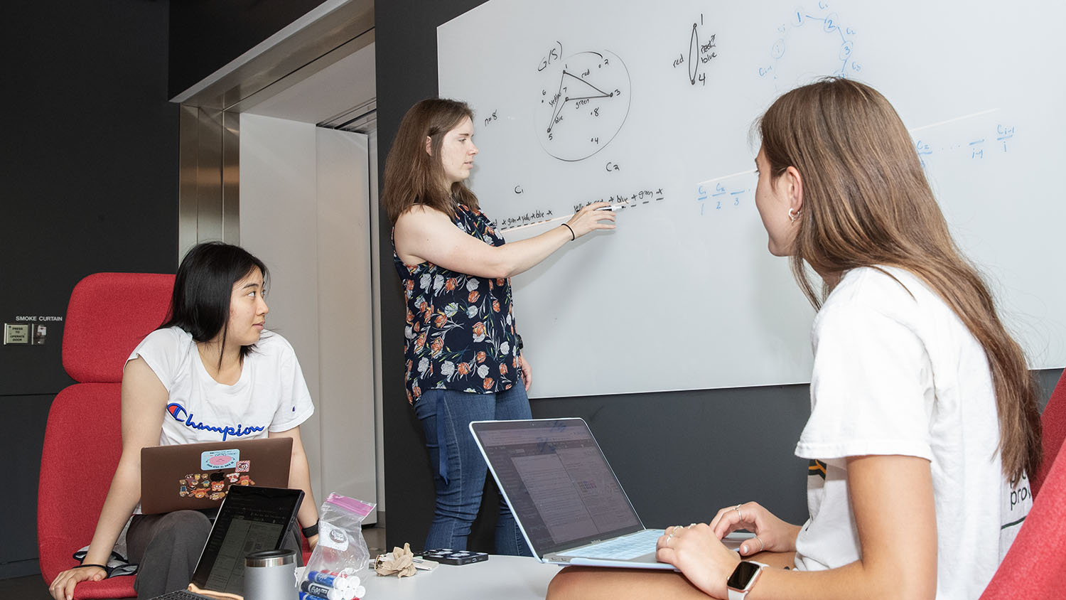 Associate Professor Kirsten Hogenson writes equations on a white board as Riley Vavolizza ’26 and Rachel Xia ’26 watch with computers on their laps.