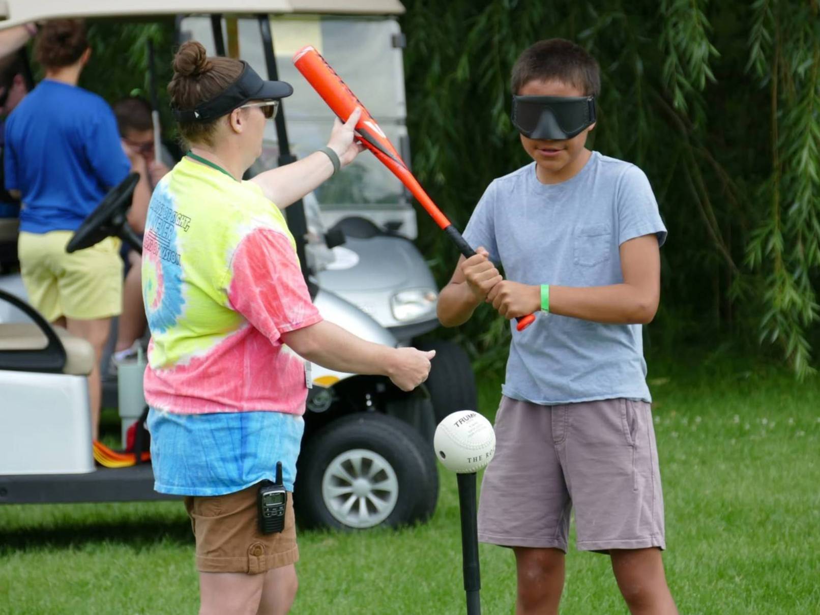A young camper plays beep baseball. They hold a bat, assisted by a counselor in a tie-dye shirt