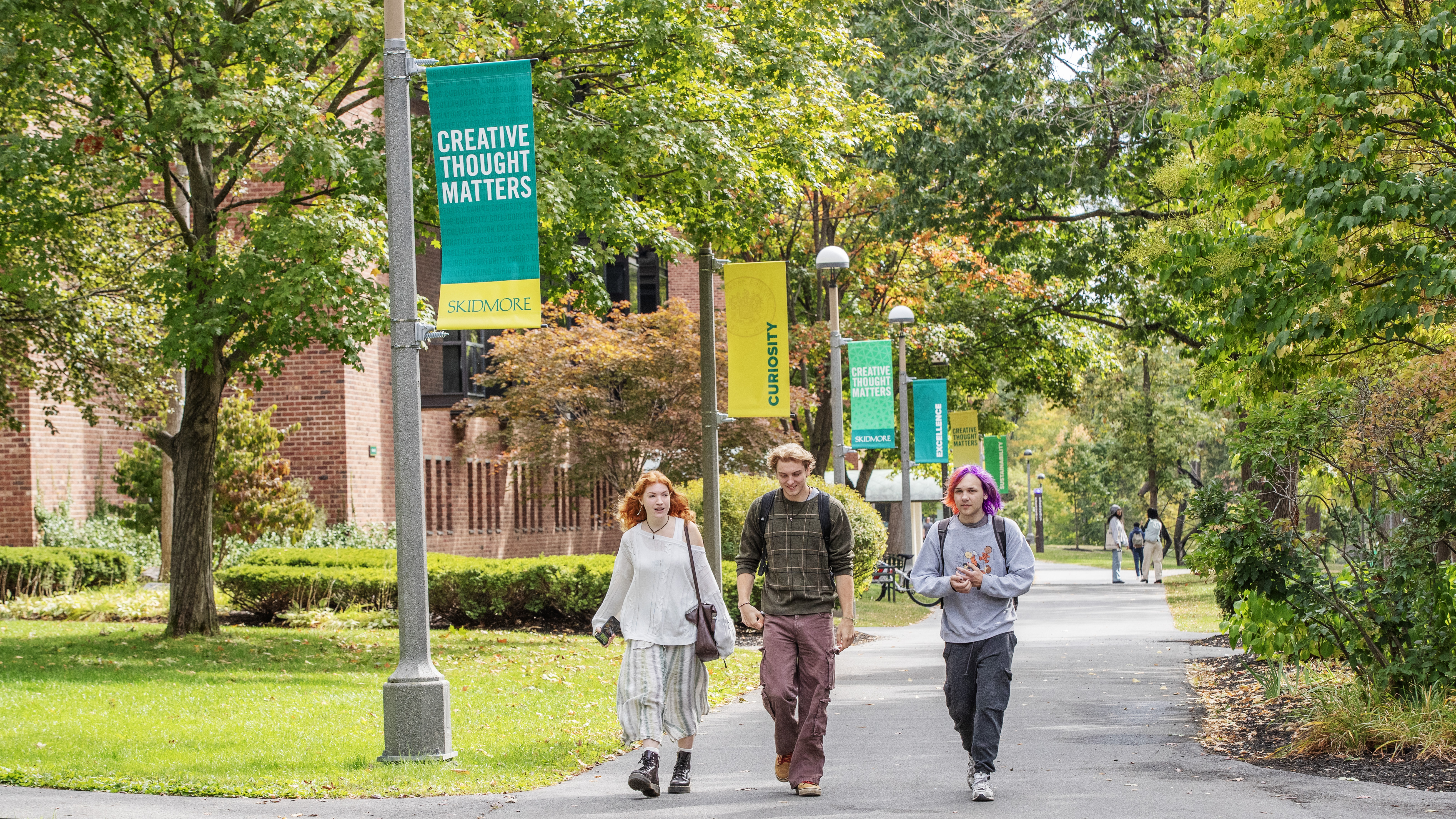 Students walk on Skidmore's campus near flags reading "Creative Thought Matters" and "curiosity." 