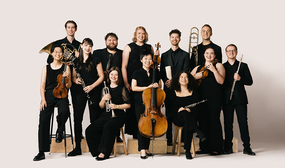 Twelve musicians in black pose with their instruments against a light background, smiling at the camera